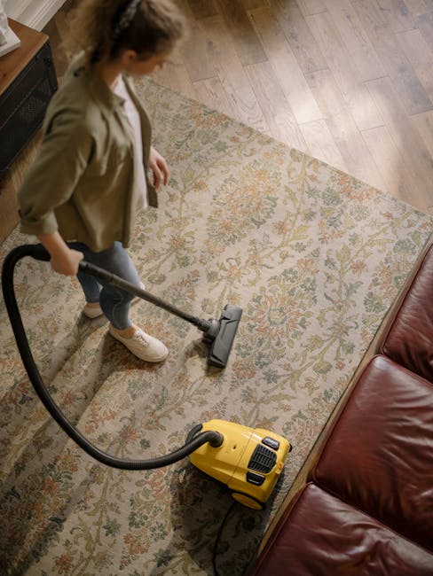 A professional cleaner dressed in a white protective suit, face mask, and orange gloves is operating a vacuum cleaner on a light-colored tiled floor in a modern, well-lit room with large windows and m